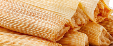 A stack of tamales in corn husks, two of them unwrapped to show the masa and red chile filling inside.